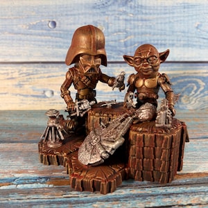 May include: A bronze-toned figurine set featuring two characters, one resembling a masked figure in a helmet and the other a small creature with pointed ears, positioned on a textured base with miniature metallic starship and accessory models.