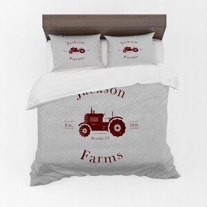 May include: A bed set featuring a gray duvet cover and two pillowcases. The set displays a maroon tractor graphic with the text "Jackson Farms" and "Riverside, VT" on the duvet. The pillowcases repeat the tractor design.