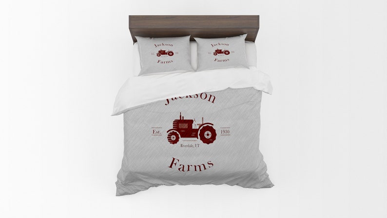 May include: A bed set featuring a gray duvet cover and two pillowcases. The set displays a maroon tractor graphic with the text "Jackson Farms" and "Riverside, VT" on the duvet. The pillowcases repeat the tractor design.