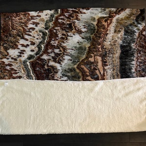 May include: A white throw blanket with a brown, green, and white patterned fleece throw blanket on top. The fleece blanket has a marbled pattern.