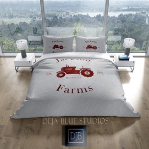 May include: A light gray bed set featuring a red tractor design with the words "Jackson Farms" and "Riverdale, UT" printed on the duvet cover and two pillowcases. The set includes a duvet cover and two pillowcases.