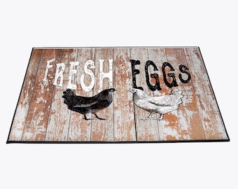May include: A brown and white doormat with a rustic woodgrain design. The text "FRESH EGGS" is printed in black with a white outline. Two black and white chickens are pictured on either side of the text.