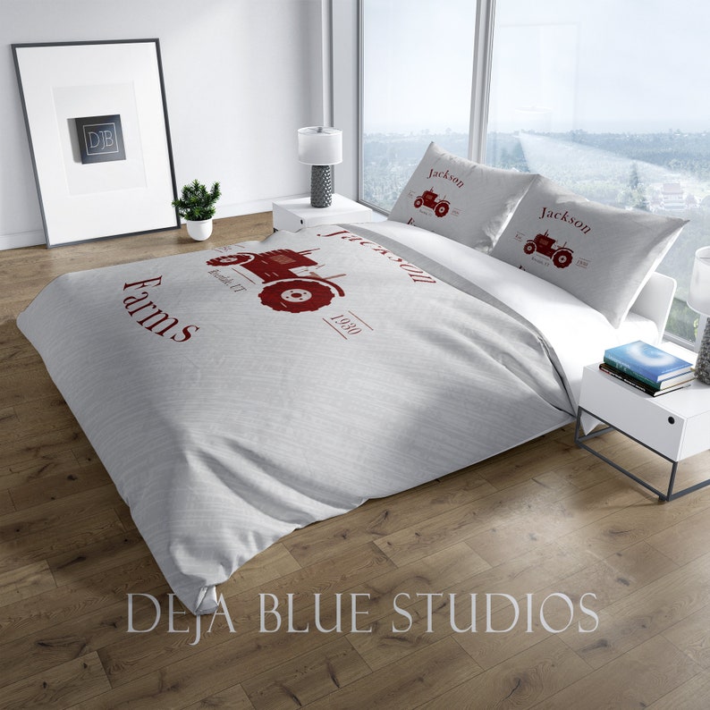 May include: A light gray bed set featuring a red tractor graphic and the text "Jackson Farms" and "1930". The set includes a duvet cover and two pillowcases. The bedding is on a bed in a well-lit room.