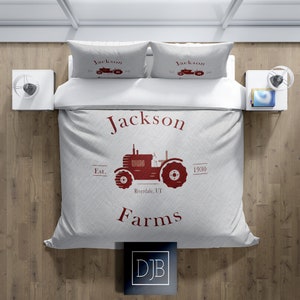 May include: A light gray bedding set featuring a red tractor design with the words "Jackson Farms" and "Est. 1930 Riverdale, UT." The set includes a duvet cover and two pillowcases. The bedding is on a white bed with two white nightstands.