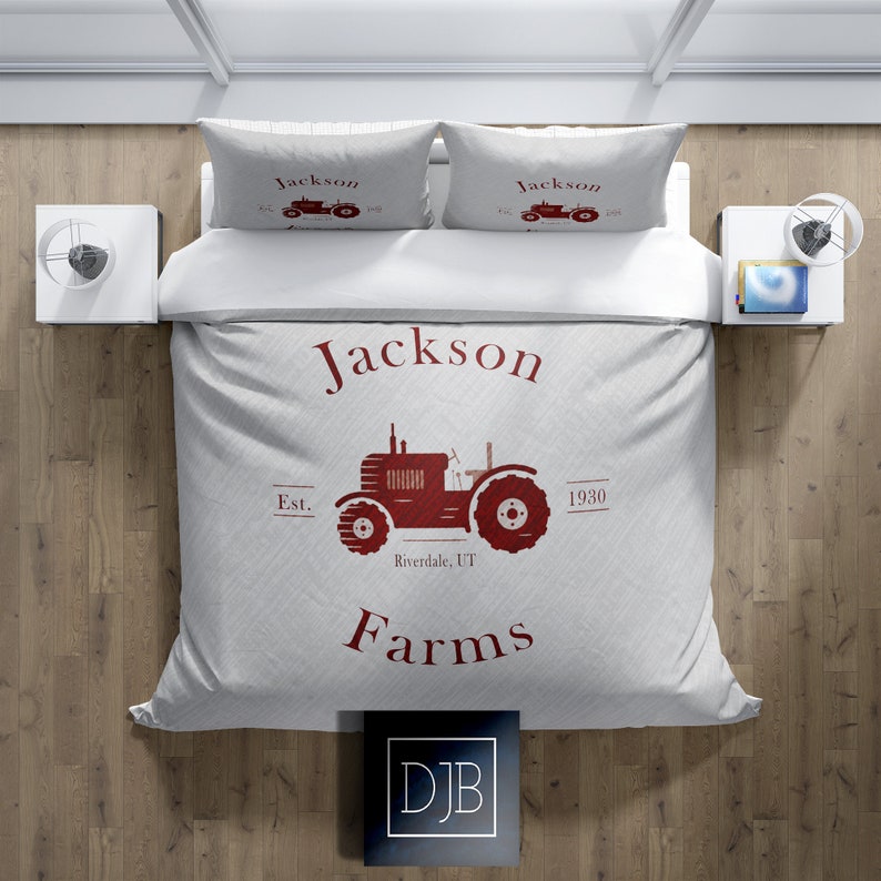 May include: A light gray bedding set featuring a red tractor design with the words "Jackson Farms" and "Est. 1930 Riverdale, UT." The set includes a duvet cover and two pillowcases. The bedding is on a white bed with two white nightstands.