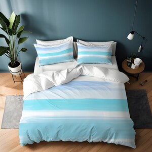 Pode incluir: Uma capa de edredom listrada azul e branca com um design semelhante à aquarela. A capa de edredom está em uma cama com fronhas brancas e um edredom branco.