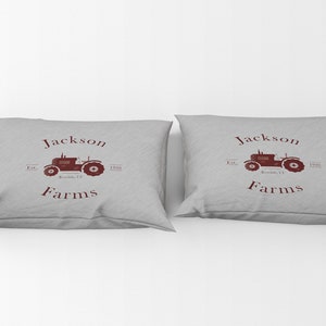 May include: Two gray throw pillows with a maroon tractor graphic and the words "Jackson Farms" printed on them. The pillows have a rectangular shape and are made of a soft fabric.