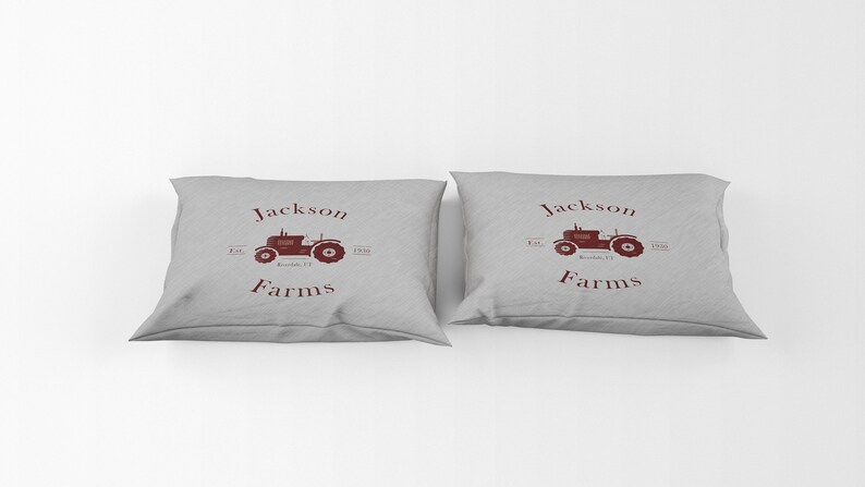 May include: Two gray throw pillows with a maroon tractor graphic and the words "Jackson Farms" printed on them. The pillows have a rectangular shape and are made of a soft fabric.
