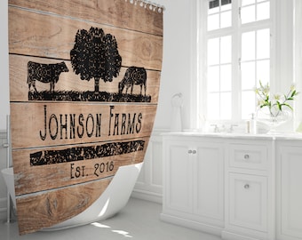 May include: A rustic shower curtain with a wood-grain design. The curtain features a black silhouette of a tree and two cows, with the text "Johnson Farms Est. 2018". The curtain is hanging in a bright white bathroom.