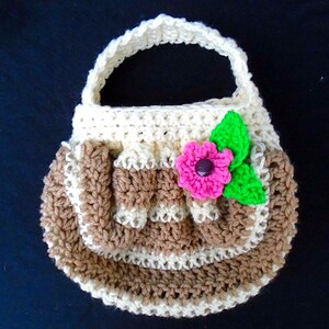 Puede incluir: Bolso de crochet hecho a mano en crema, marrón y rosa. El bolso tiene un asa y está decorado con una flor rosa con centro morado y hojas verdes. El bolso es redondo y tiene una solapa.
