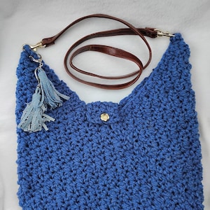 Peut inclure: Un sac bleu crocheté à la main avec une bandoulière en cuir marron et des accessoires dorés. Le sac est doté d'une fermeture à rabat avec un bouton doré et un pompon bleu décoratif. Le sac est conçu avec un point texturé.