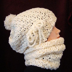 以下が含まれることがあります： クリーム色の手編み帽子とスヌードのセット。帽子にはポンポンと装飾的なリボンが付いています。スヌードは太いループのデザインです。セットは柔らかく、質感のある糸でできており、寒い季節に最適です。マネキンに着けています。