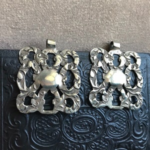 May include: Two silver-toned metal clasps with intricate floral designs. The clasps are square-shaped with a raised circular center and a loop at the top.