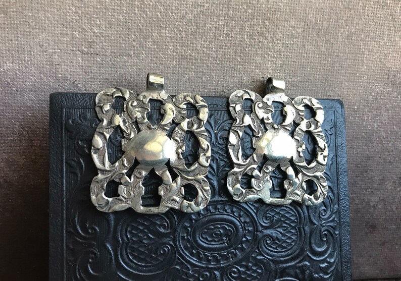 May include: Two silver-toned metal clasps with intricate floral designs. The clasps are square-shaped with a raised circular center and a loop at the top.