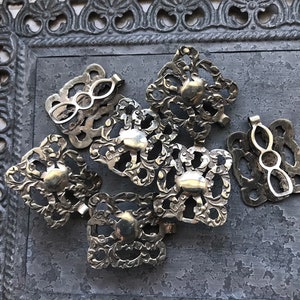 May include: A group of eight silver-toned metal clasps with intricate floral designs. The clasps are arranged in a cluster on a gray textured surface.