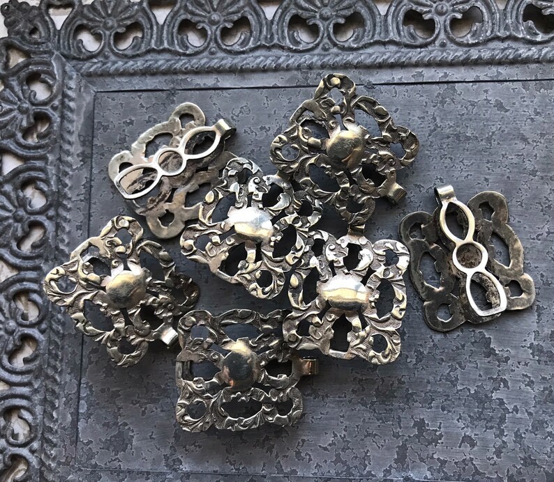 May include: A group of eight silver-toned metal clasps with intricate floral designs. The clasps are arranged in a cluster on a gray textured surface.