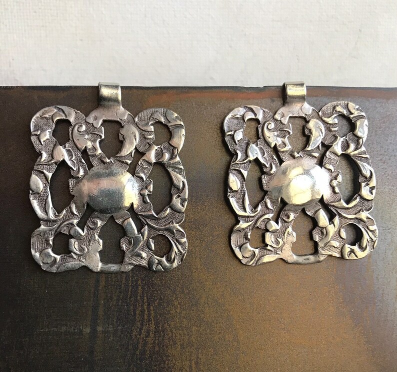 May include: Two silver, ornate, square-shaped pendants with a floral design. The pendants have a loop at the top for hanging.
