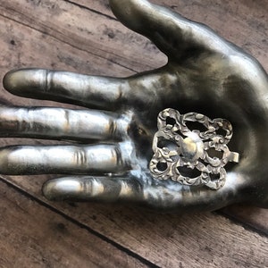 May include: A silver filigree design with a circular center, resembling a flower or a decorative element. The design is displayed on a silver hand.