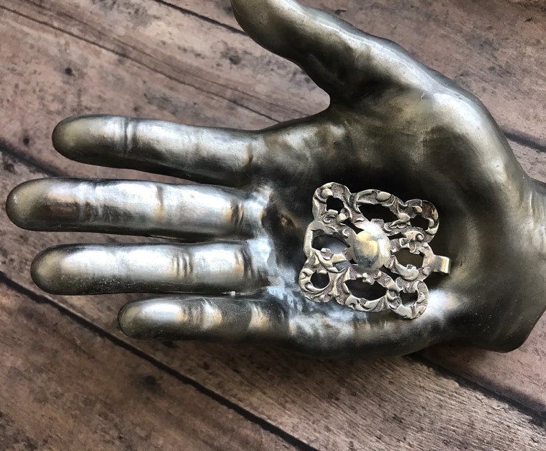 May include: A silver filigree design with a circular center, resembling a flower or a decorative element. The design is displayed on a silver hand.