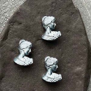 May include: Three silver-toned metal busts of women with detailed hair and clothing. The busts are arranged in a triangular pattern on a dark gray textured surface.