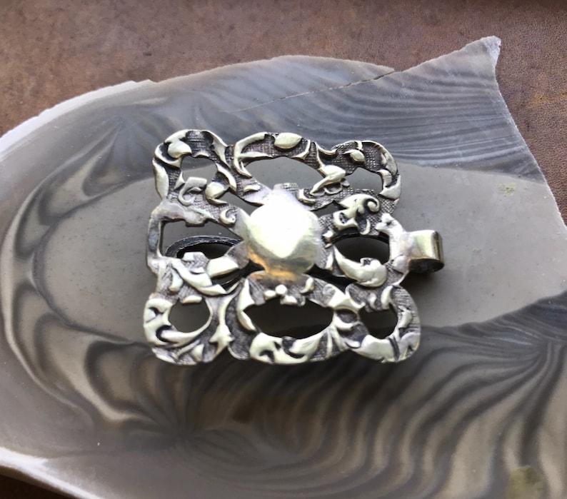 May include: A silver, ornate, decorative buckle with a floral design.