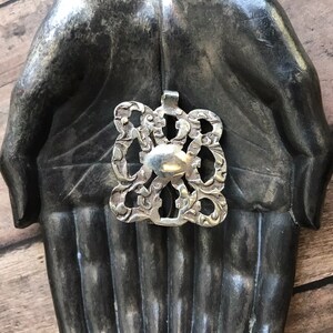 May include: A silver pendant with an intricate design, featuring a central oval shape surrounded by swirling patterns. The pendant is displayed on a pair of hands sculpted from a dark gray material.