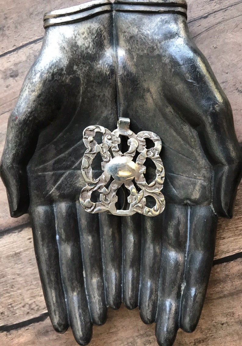 May include: A silver pendant with an intricate design, featuring a central oval shape surrounded by swirling patterns. The pendant is displayed on a pair of hands sculpted from a dark gray material.