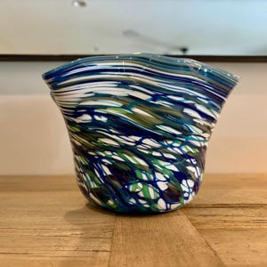 May include: A hand-blown glass vase with a flared, wavy rim. The vase features a swirling pattern of blue, white, and green colors. The glass is thick and has a textured appearance, creating a unique decorative piece.