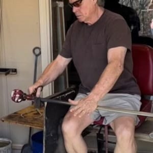 Puede incluir: Una persona trabajando con vidrio, sosteniendo una herramienta de soplado de vidrio. La persona lleva una camisa marr&oacute;n, pantalones cortos grises y gafas de sol. La persona est&aacute; sentada cerca de una mesa con herramientas y una silla roja.