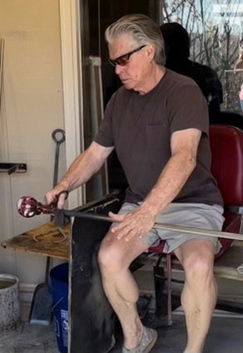 Puede incluir: Una persona trabajando con vidrio, sosteniendo una herramienta de soplado de vidrio. La persona lleva una camisa marr&oacute;n, pantalones cortos grises y gafas de sol. La persona est&aacute; sentada cerca de una mesa con herramientas y una silla roja.