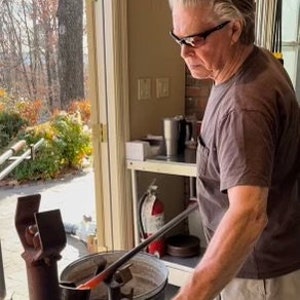 Puede incluir: Un artesano trabajando con vidrio fundido, utilizando una varilla met&aacute;lica para dar forma al material incandescente. La escena se desarrolla en un taller con luz natural, con un extintor y varias herramientas.