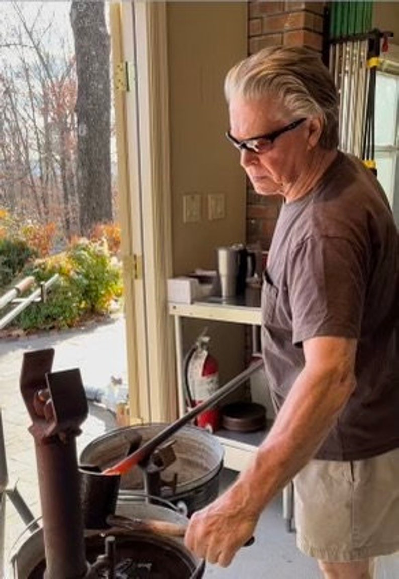 Puede incluir: Un artesano trabajando con vidrio fundido, utilizando una varilla met&aacute;lica para dar forma al material incandescente. La escena se desarrolla en un taller con luz natural, con un extintor y varias herramientas.