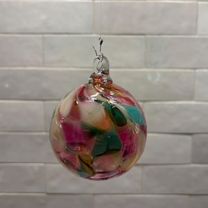 May include: A spherical glass ornament featuring a swirling pattern of pink, teal, white, and amber colors. The ornament has a clear glass loop at the top with a small metal hook attached, allowing it to hang from a thin, nearly invisible string.