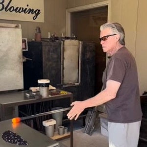 Puede incluir: Un soplador de vidrio en un taller, con gafas de sol y una camiseta marr&oacute;n, trabajando con vidrio fundido. La palabra "Blowing" es visible en un letrero. Una mesa con herramientas y materiales est&aacute; en primer plano.