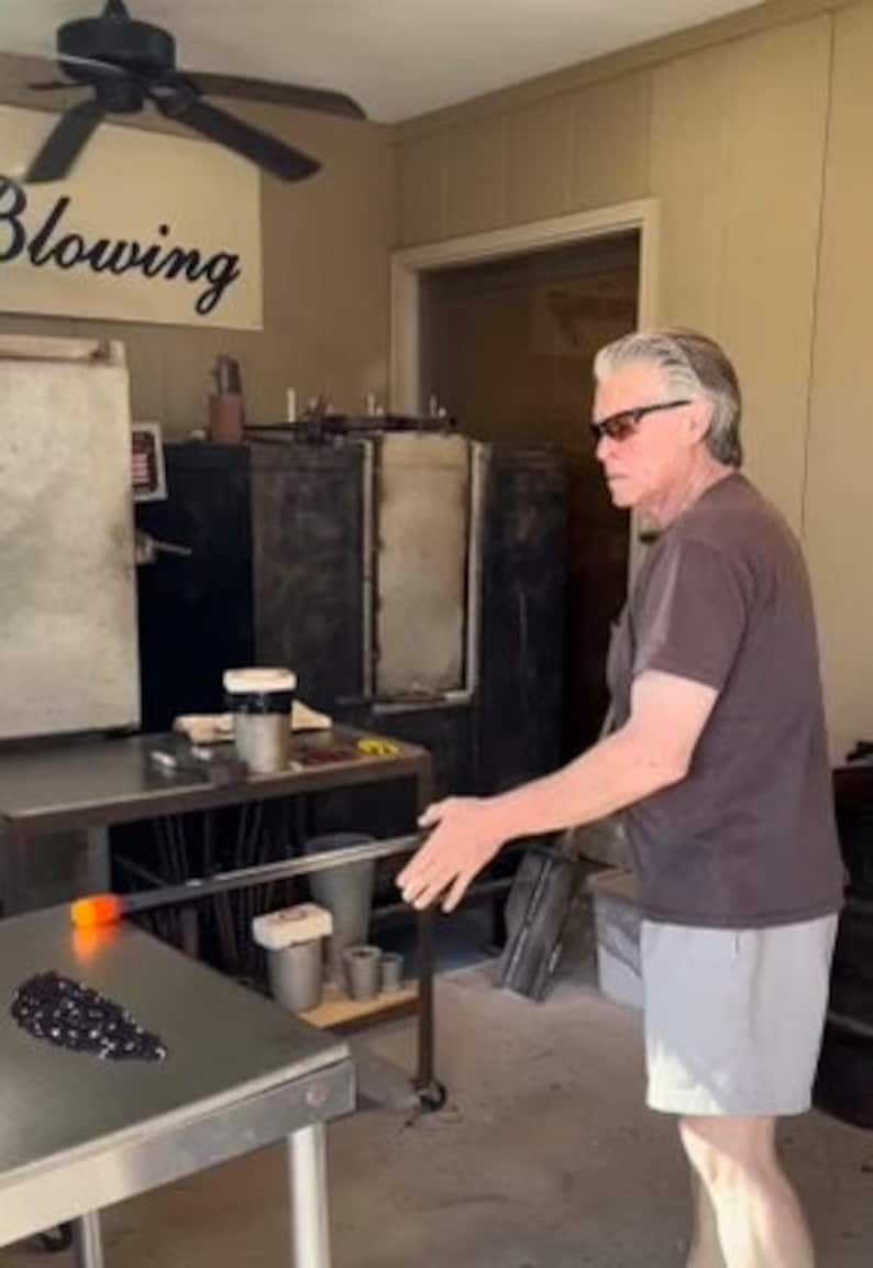 Puede incluir: Un soplador de vidrio en un taller, con gafas de sol y una camiseta marr&oacute;n, trabajando con vidrio fundido. La palabra "Blowing" es visible en un letrero. Una mesa con herramientas y materiales est&aacute; en primer plano.