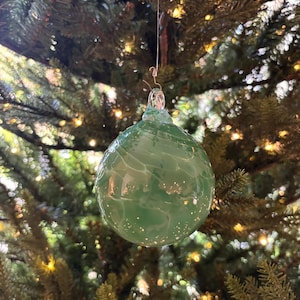 May include: A green glass Christmas ornament with a swirled pattern and a silver wire hanger. The ornament is round and has a glossy finish. It is hanging on a Christmas tree with green needles and small lights.