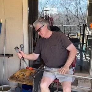 Puede incluir: Una persona trabajando con herramientas de soplado de vidrio, vistiendo una camiseta marr&oacute;n y pantalones cortos. La persona sostiene una varilla de metal y manipula vidrio fundido. El espacio de trabajo incluye una peque&ntilde;a mesa, un cubo y una ventana con vista a &aacute;rboles.