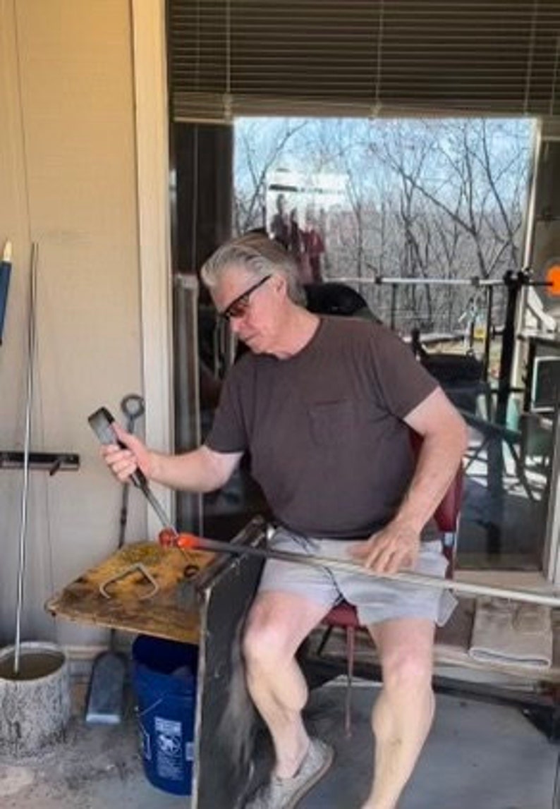 Puede incluir: Una persona trabajando con herramientas de soplado de vidrio, vistiendo una camiseta marr&oacute;n y pantalones cortos. La persona sostiene una varilla de metal y manipula vidrio fundido. El espacio de trabajo incluye una peque&ntilde;a mesa, un cubo y una ventana con vista a &aacute;rboles.