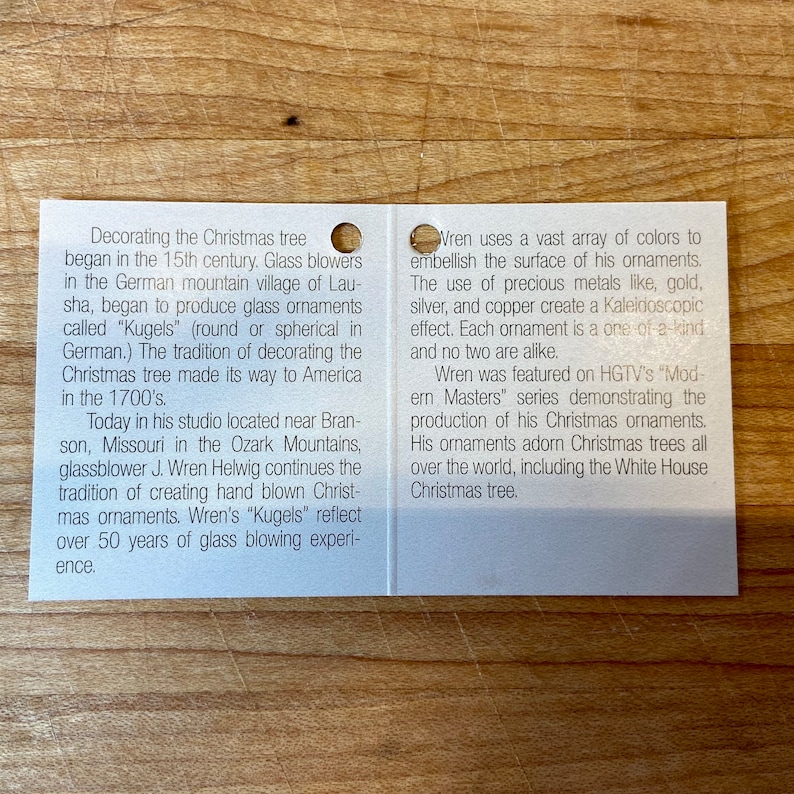 Puede incluir: Una tarjeta blanca con texto sobre adornos navide&ntilde;os, incluyendo la historia del soplado de vidrio y el uso de metales preciosos. La tarjeta menciona al soplador de vidrio J. Wren Helwig y su estudio en Misuri.
