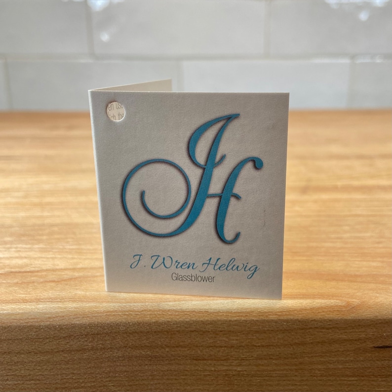 Puede incluir: Una tarjeta plegada con las iniciales turquesas "JH" en una fuente decorativa. La tarjeta tambi&eacute;n presenta el texto "J. Wren Helwig Glassblower" en una fuente m&aacute;s peque&ntilde;a y sencilla. La tarjeta est&aacute; sobre una superficie de madera.