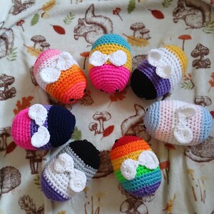 May include: Seven colourful, crocheted egg-shaped objects, each with a white bow. The eggs feature rainbow, pink, blue, and black colour schemes. The objects are arranged on a patterned fabric with woodland creatures.