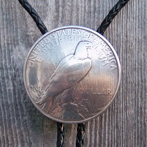 May include: A silver Peace Dollar coin bolo tie with a black braided leather cord. The coin features an image of a bald eagle with outstretched wings and the words "United States of America" and "One Dollar" around the edge.