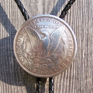 May include: A silver dollar coin bolo tie with a bald eagle design and the inscription "United States of America, In God We Trust, One Dollar".