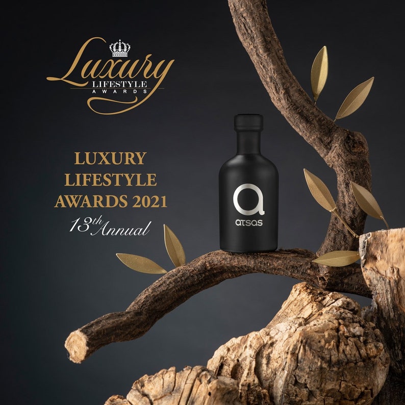 Puede incluir: Una botella de vidrio negro con una etiqueta plateada que dice "atsas" y una gran letra plateada "a" en la parte delantera. La botella est&aacute; sentada en una rama de un &aacute;rbol con hojas doradas. El texto "Luxury Lifestyle Awards 2021 13th Annual" est&aacute; en el fondo.