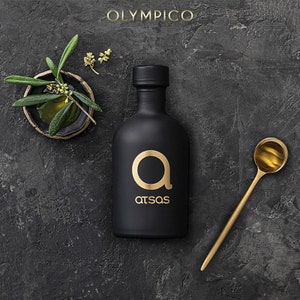 K&ouml;nnte beinhalten: Eine schwarze Oliven&ouml;lflasche mit einem goldenen "a"-Logo und dem Wort "atsas" in Gold. Ein goldener L&ouml;ffel und eine kleine Schale mit Olivenzweigen sind ebenfalls vorhanden. Das Wort "OLYMPICO" steht oben. Der Hintergrund ist dunkelgrau.