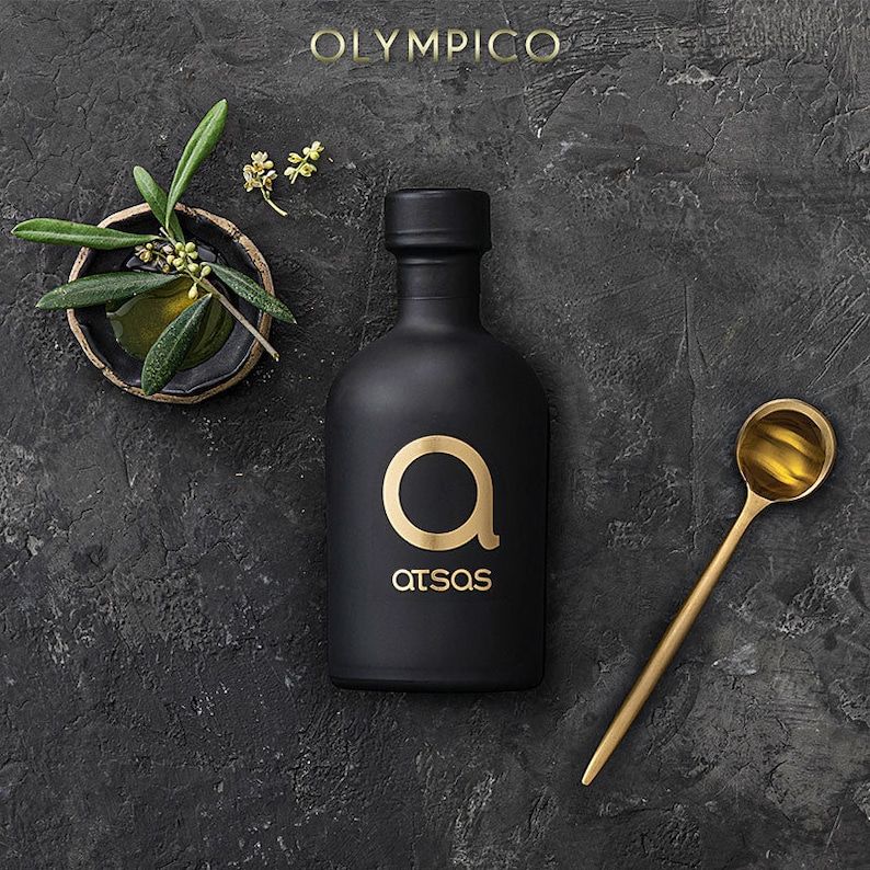 K&ouml;nnte beinhalten: Eine schwarze Oliven&ouml;lflasche mit einem goldenen "a"-Logo und dem Wort "atsas" in Gold. Ein goldener L&ouml;ffel und eine kleine Schale mit Olivenzweigen sind ebenfalls vorhanden. Das Wort "OLYMPICO" steht oben. Der Hintergrund ist dunkelgrau.