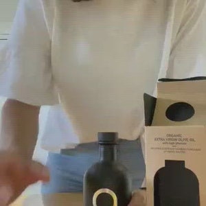 Puede incluir: Una botella de vidrio negro de aceite de oliva virgen extra org&aacute;nico con una etiqueta dorada que dice "atsos" y una letra dorada "A" dentro de un c&iacute;rculo. La botella est&aacute; sentada sobre una mesa de madera junto a una caja con un dise&ntilde;o similar.