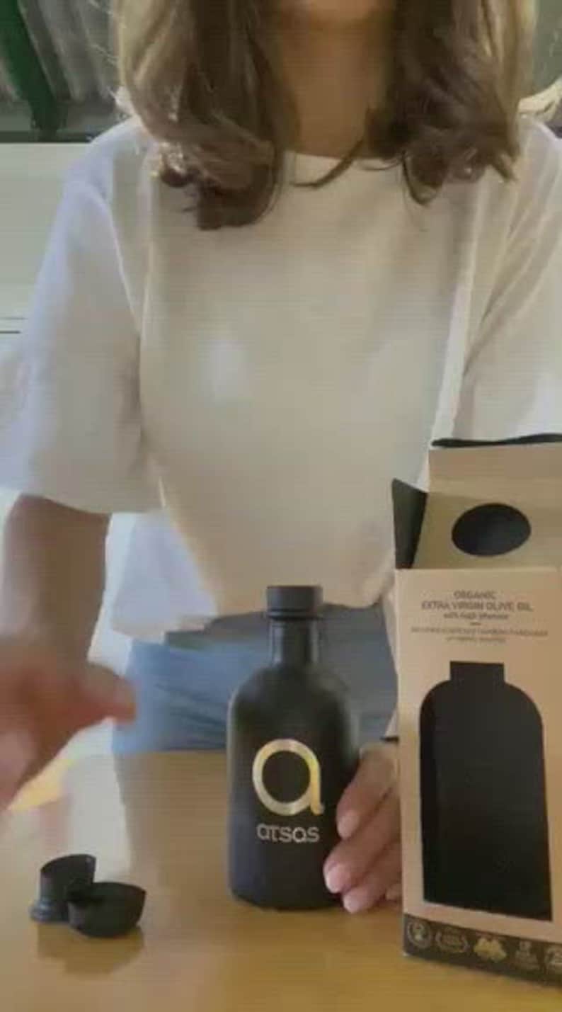 Puede incluir: Una botella de vidrio negro de aceite de oliva virgen extra org&aacute;nico con una etiqueta dorada que dice "atsos" y una letra dorada "A" dentro de un c&iacute;rculo. La botella est&aacute; sentada sobre una mesa de madera junto a una caja con un dise&ntilde;o similar.