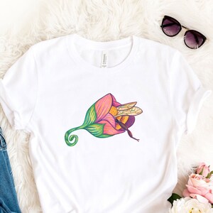 Può includere: Maglietta bianca con un'illustrazione colorata di un'ape all'interno di un fiore. La maglietta è stesa su una superficie bianca e soffice con un paio di occhiali da sole, un paio di scarpe da ginnastica bianche e un mazzo di fiori rosa e bianchi.