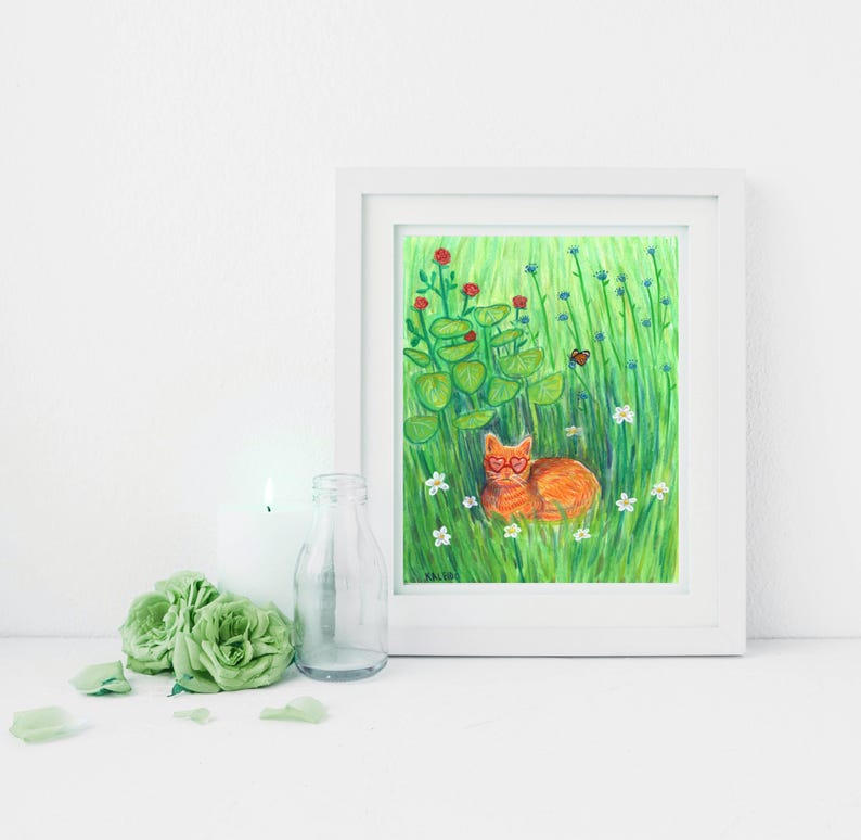 K&ouml;nnte beinhalten: Ein gerahmter Kunstdruck mit einer orangefarbenen Katze mit herzf&ouml;rmiger Brille, die in einem Feld aus gr&uuml;nem Gras, Blumen und Bl&auml;ttern liegt. Das Kunstwerk befindet sich in einem wei&szlig;en Rahmen mit wei&szlig;em Hintergrund. Eine Kerze, eine Glasflasche und gr&uuml;ne Rosen befinden sich im Vordergrund.